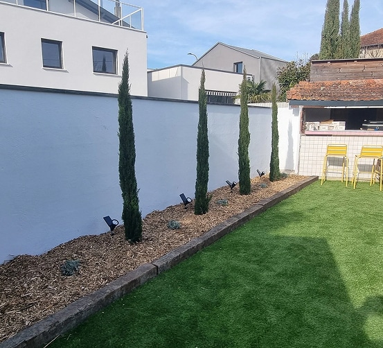 Jardin moderne avec pelouse artificielle, quatre cyprès élancés bordant un mur blanc et des spots lumineux orientés vers le haut.