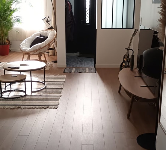 Salon lumineux avec fauteuil rotin blanc, tables gigognes, tapis rayé, guitare électrique contre le mur près de l'entrée noire.