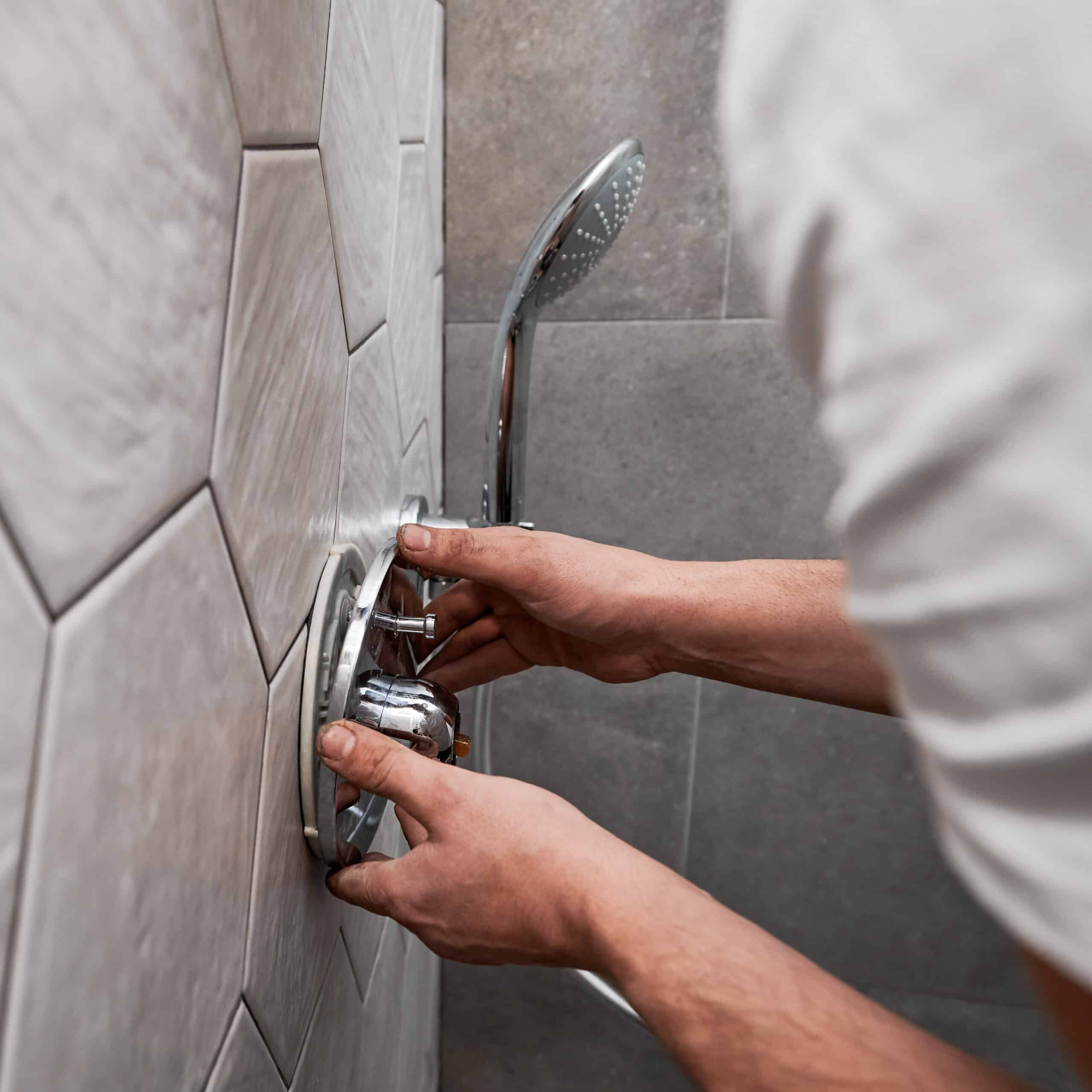 Plombier installant une poignée chromée de mitigeur de douche sur un mur de carreaux hexagonaux gris et beiges.